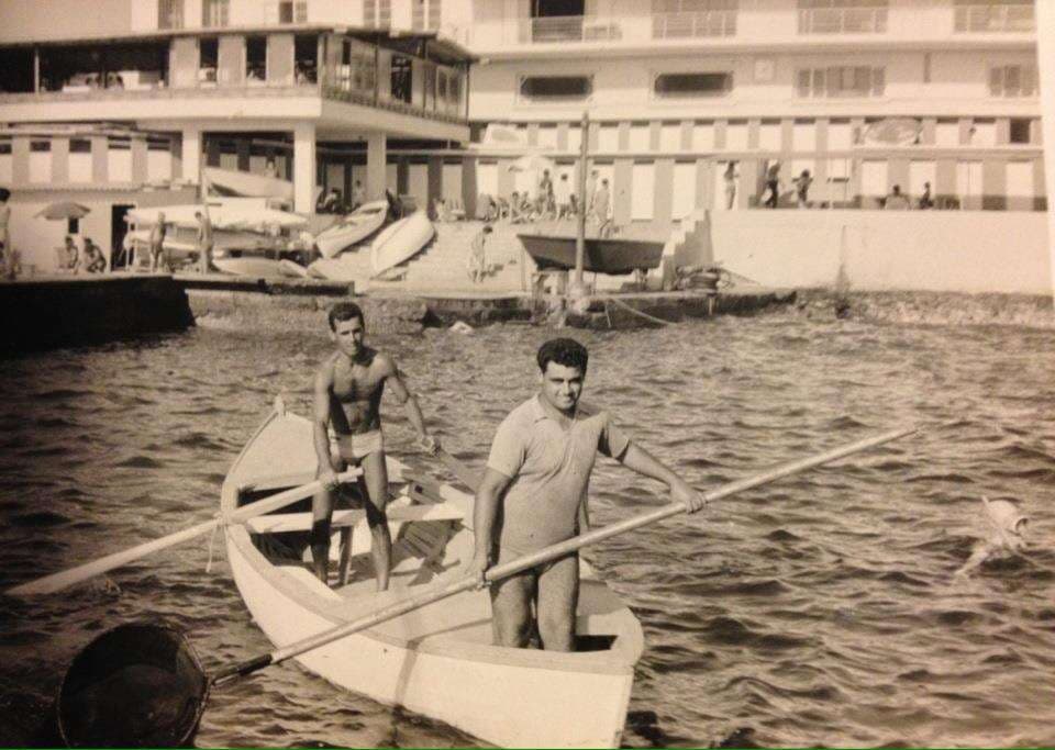 Two men in a rowboat off Sporting Club Beach, the clubhouse behind — an archive photograph