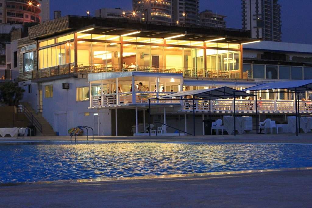 The waterfront restaurants at Sporting Club Beach lit at dusk, seen across the pools
