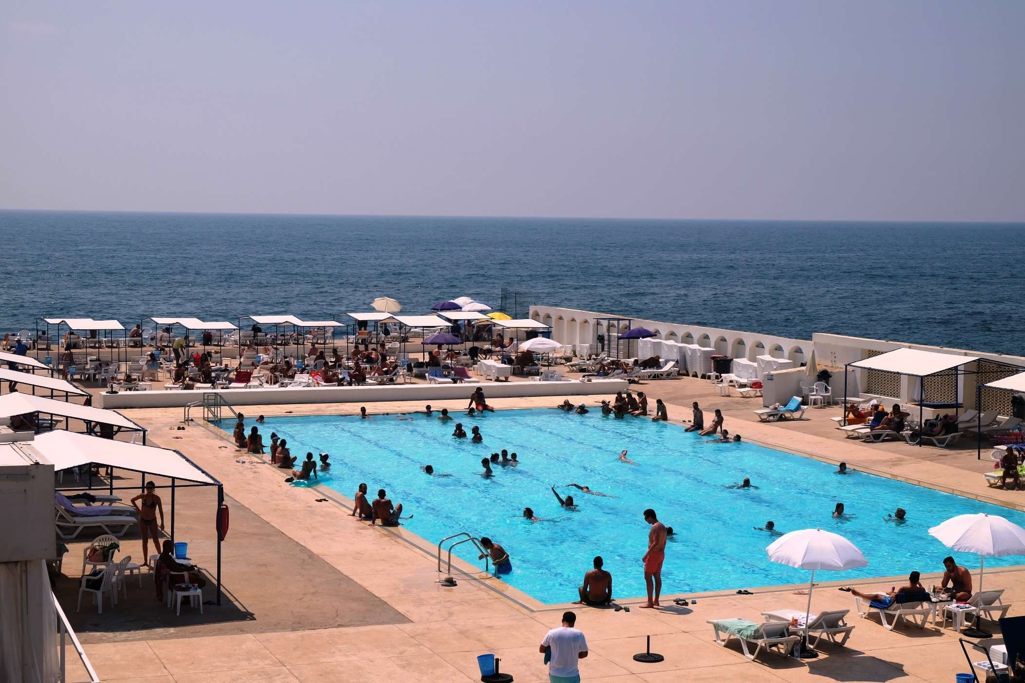 A busy saltwater pool ringed by umbrellas and arched cabanas at Sporting Club Beach, Ras Beirut