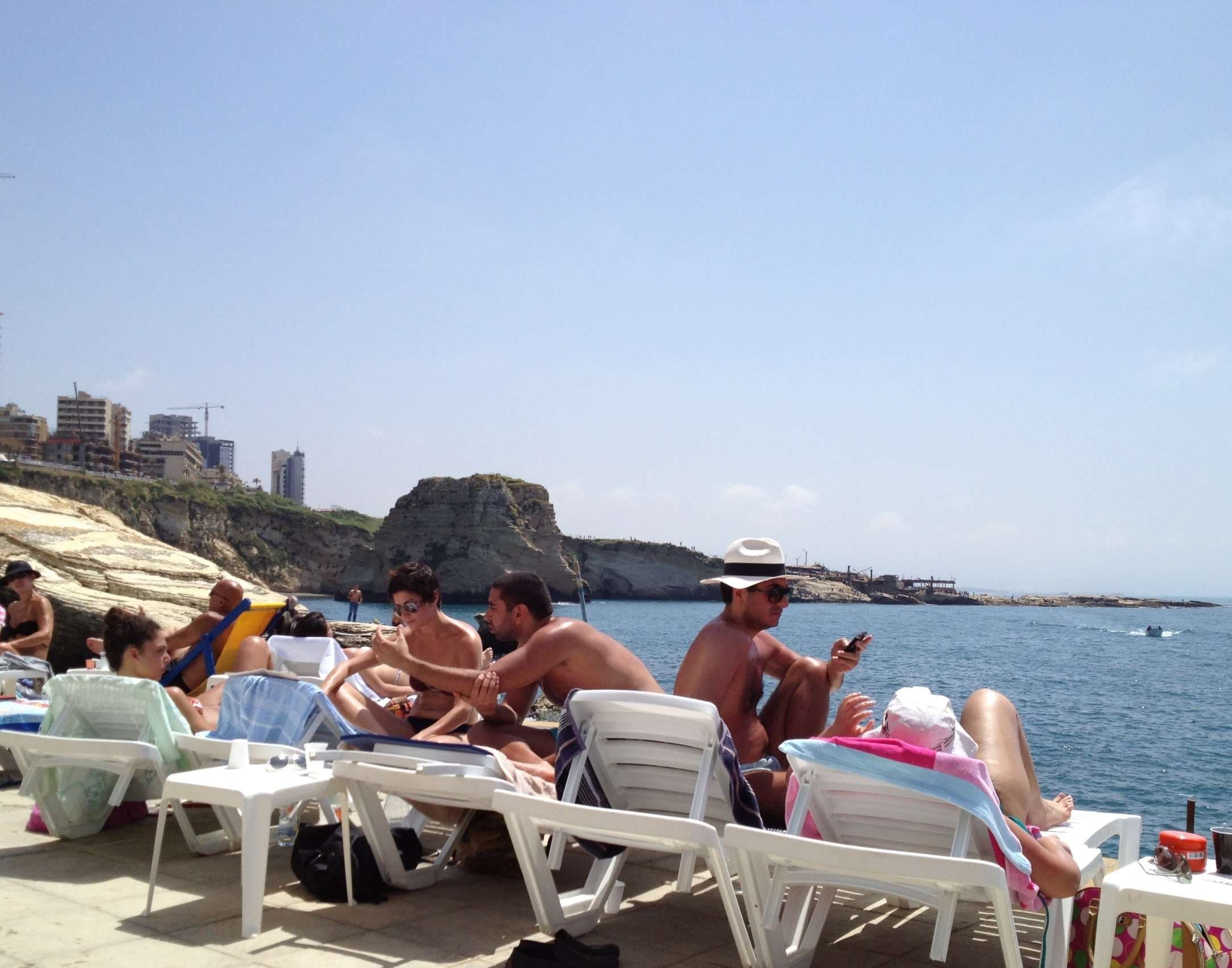 Friends on sun loungers along the rocky shoreline with the Pigeon Rocks behind at Sporting Club Beach, Ras Beirut