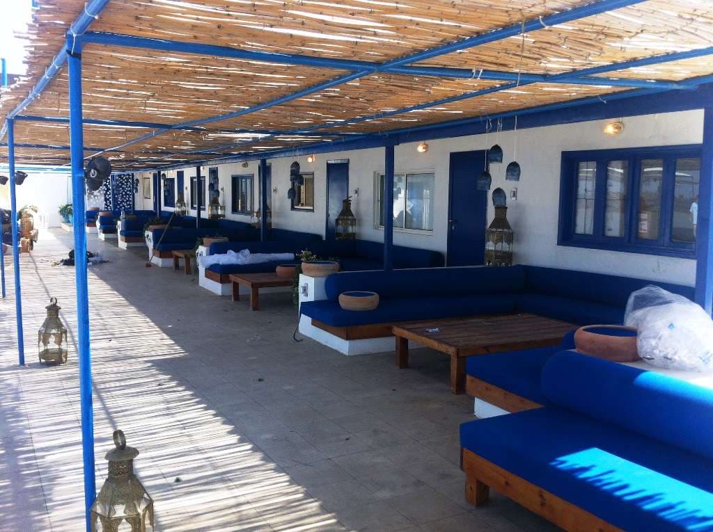 A covered seaside lounge lined with blue banquettes and brass lanterns at Sporting Club Beach