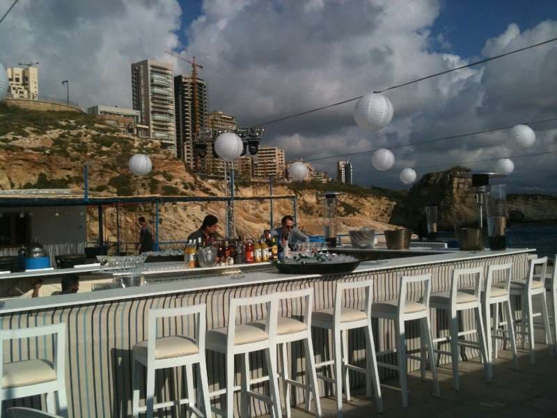 A seafront bar with white stools and lanterns against the Ras Beirut cliffs at Sporting Club Beach