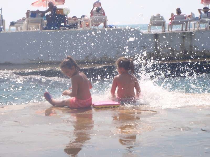 Children playing in the shallows as waves splash over the deck at Sporting Club Beach, Ras Beirut