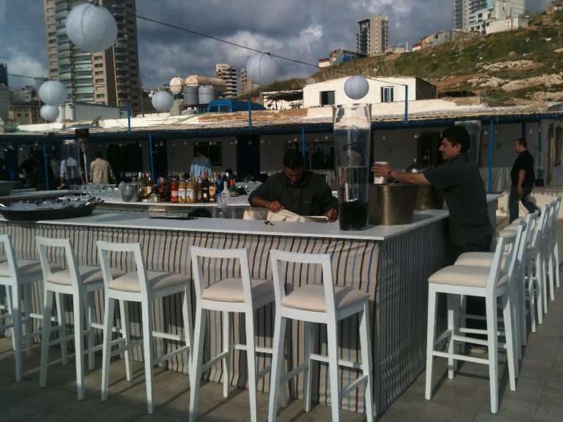 An outdoor bar counter with a bartender, the coastline behind, at Sporting Club Beach